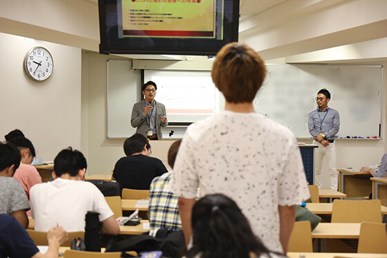 大学の教室で、マイクを手に学生へ向けて話す吉川健太