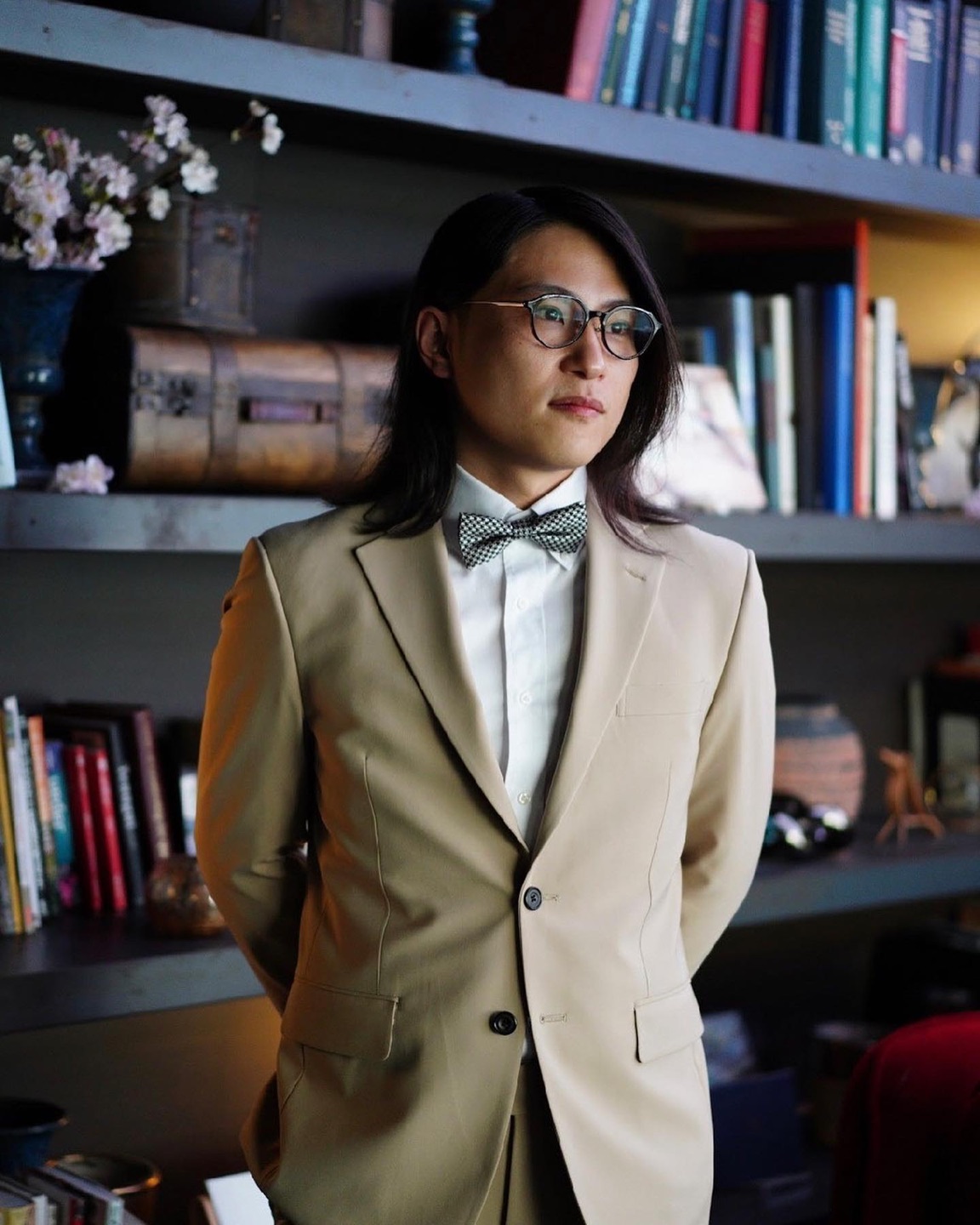 Kenta Yoshikawa in a tailored beige suit, bow tie, and round glasses, photographed in front of bookshelves.